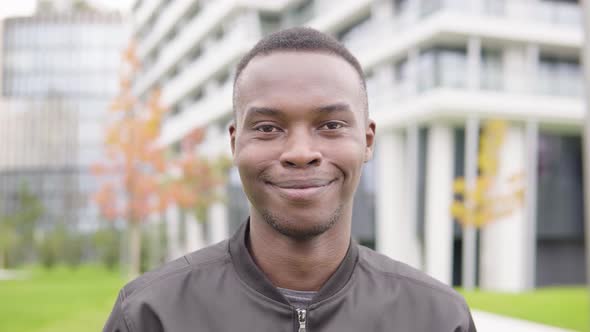 A Young Black Man Smiles at the Camera - Closeup - Office Buildings in the Blurry Background