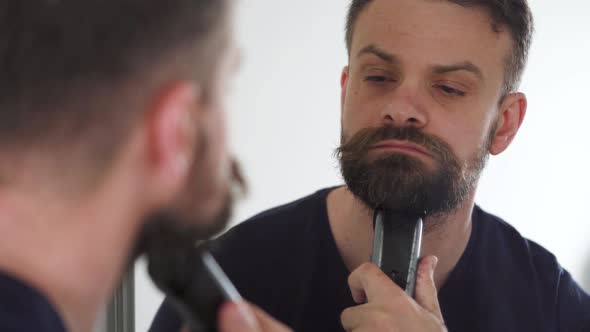 Man Shaves His Beard Using an Electric Trimmer. Reflection in the Mirror