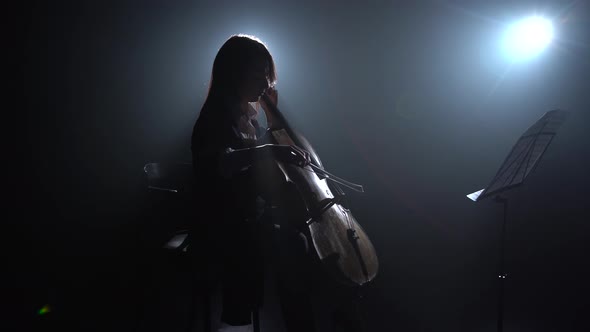 Musician Girl in a Dark Room Rehearsing a Cello Playing and Rubbed Into a Music Stand. Silhouette