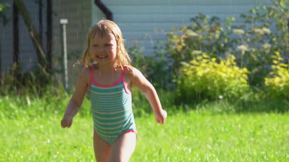 The Cute Little Girl Is Running Around a Revolving Water Sprinkler on the Lawn