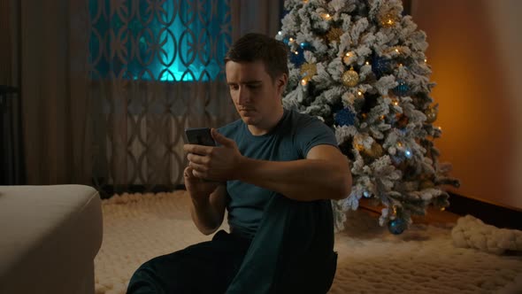 Young Caucasian Man Using Smartphone Sitting Near Cozy Christmas Tree Space