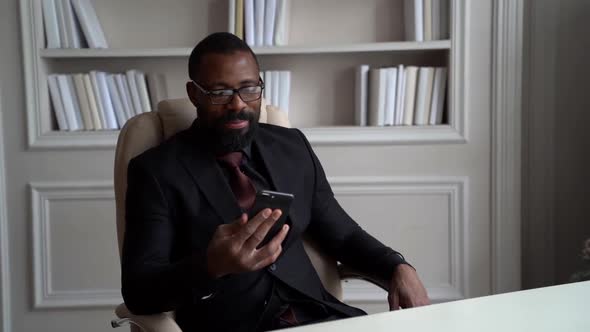 a Bearded AfricanAmerican Man in Glasses and a Suit with a Tie is Sitting on a Light Chair at a Desk
