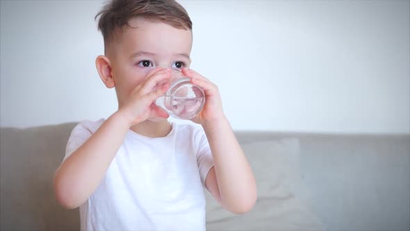 Cute Baby Boy Drinking a Glass of Water Sitting on the Couch at Home. Slow Motion Little Boy