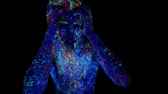 Young man in glowing neon bodyart holding his head in pain, panting, screaming in darkness. Concept