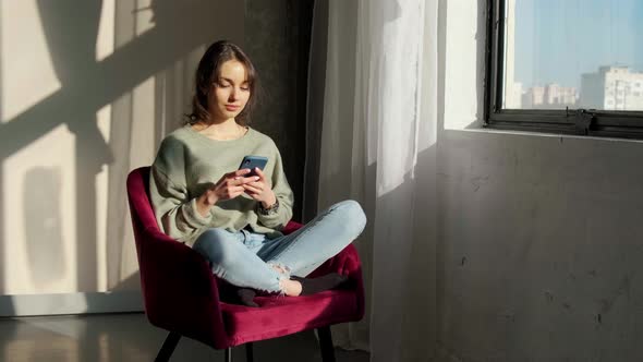 Beautiful Woman Sitting Near Window and Using Mobile Phone at Sunrise