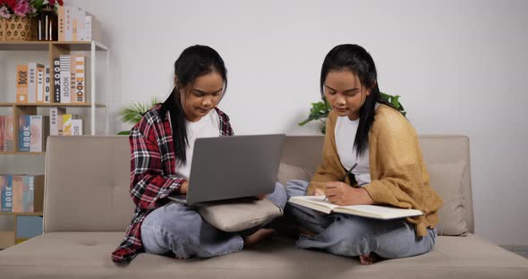 Twin girls reading a book and looking laptop while sitting on couch