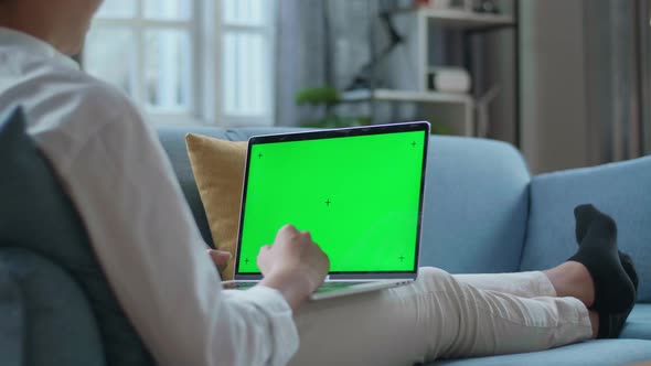 Man Waving Hand And Talking To Green Screen Laptop While Lying On Sofa In The Living Room