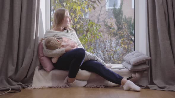 Portrait of Young Happy Caucasian Woman Sitting on Low Windowsill and Holding Her Daughter on Hands