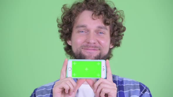 Face of Happy Young Bearded Hipster Man Thinking While Showing Phone