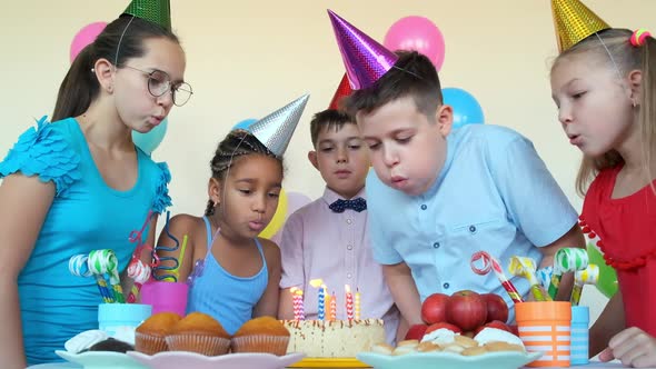 Children with Party Cones Blow Out Burning Candles on Cake