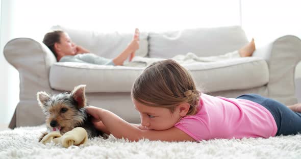 Little Girl Playing with Puppy Chewing Bone with her Mother Reading on the Sofa