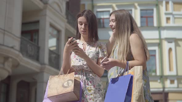 Two Adorable Happy Girlfriends After Shopping with Shopping Bags Texting on the Cellphone in Front