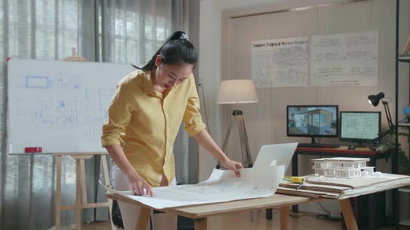 Asian Woman Engineer With A Laptop Looking At Blueprint While Working At The Office