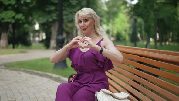 Zoom in to Happy Blond Smiling Woman with Hazel Eyes Gesturing Heart Shape with Hands Posing on