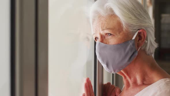 Senior caucasian woman wearing face mask looking out of window at home