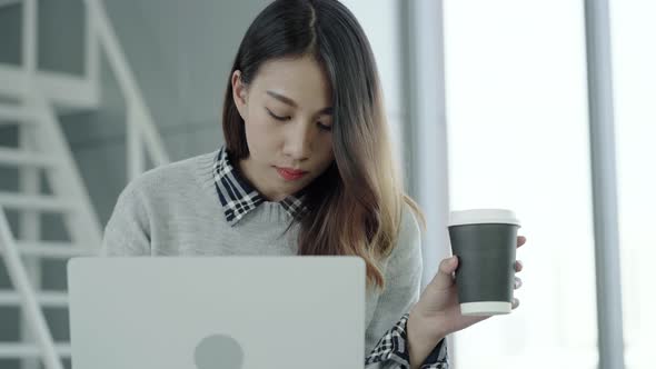 Young Asian female manager using portable computer device and drinking coffee cup at office