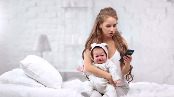 Caring Young Mother Calming Down Little Crying Baby During Using Mobile Phone Medium Shot