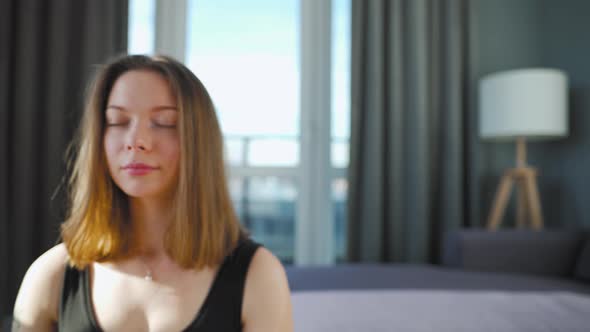 Young Caucasian Woman in Black Jumpsuit Sitting in Lotus Position and Meditating Yoga at Home