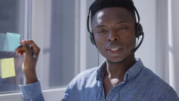 Young Inspired African American Man Writing on Sticky Notes on Window Talking in Earphones