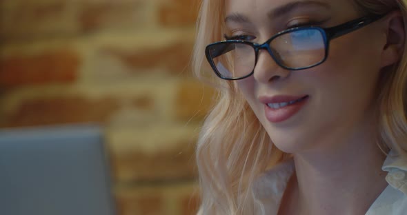 Closeup View of a Smiling Blonde Woman Wearing Eyeglasses is Working on Her Laptop in the Cafe