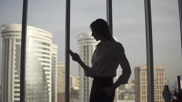 Silhouette of a Young Woman Against the Background of a Large Window and Skyscraper. Girl Uses a