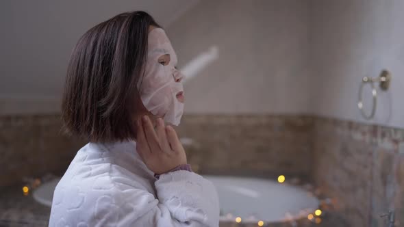 Side View Young Little Woman in Moisturizing Facial Mask Walking to Mirror in Bathroom