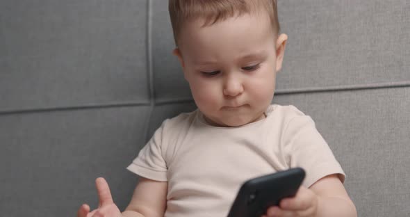 Beautiful Little Baby Boy Holds Smartphone in Hands and Looks at It Sitting on Bed