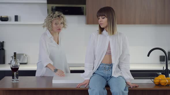 Sad Young Lesbian Woman Sitting on Countertop As Angry Partner Shouting Drinking Wine at Background