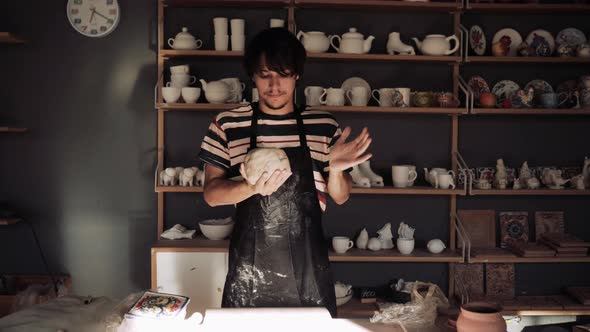 Young Male Potter Kneads Clay Against Background of a Rack with Ceramic Blanks