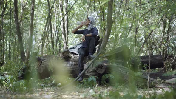 Wide Shot Portrait of Panicked Nervous Young Man Sitting in Summer Forest Drinking Water and Eating