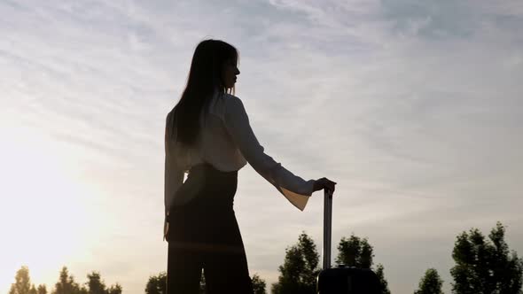 Woman Silhouette Stands with Suitcase at Back Cloudy Sunset
