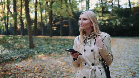 Young Girl Walking in Autumn Park and Texting a Message on a Mobile Phone