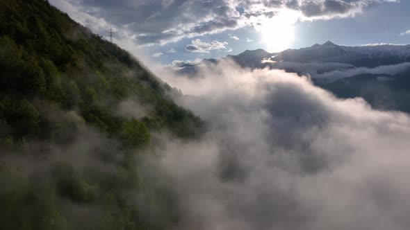 Flight above gray puffy clouds enveloping dark forested alp hillside in sunlight