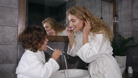 Woman with Long Curly Hair in White Bathrobe Brushing Teeth with Her Cute Son in the Morning