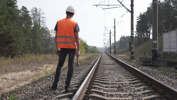 The railway worker stands along the railway tracks, holding a sledgehammer in his hands.