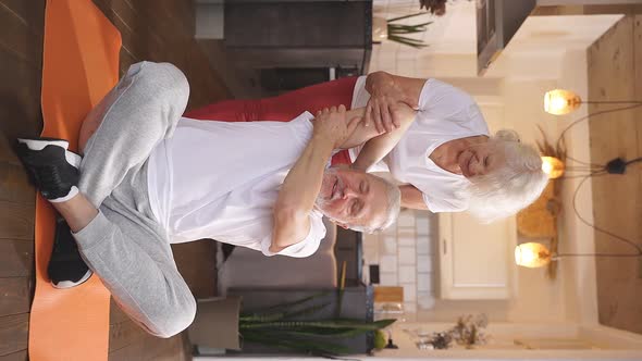An Older Couple Does Yoga and Fitness at Home a Woman Helps a Man Do Muscle Stretching While Sitting