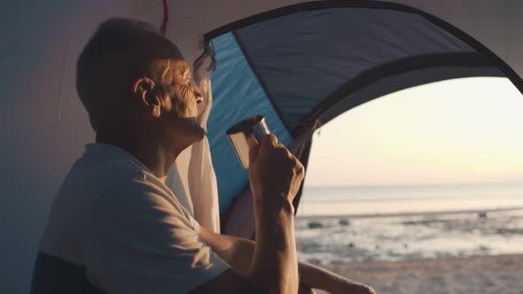 Mature Man in Tent Drinking Tea From Steel Cup with Seaside Background.