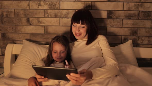 Mother and Little Preschool Daughter Lying Under Blanket on Bed Watching Video on Tablet