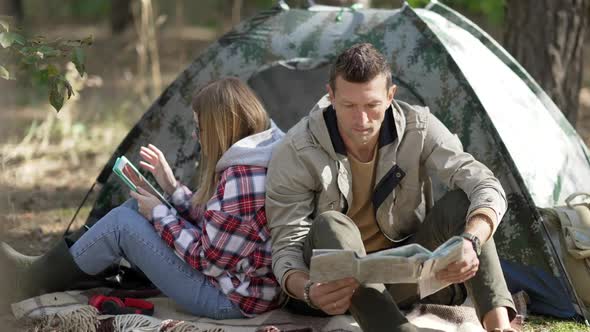 Portrait of Concentrated Man Examining Paper Map As Relaxed Woman Surfing Social Media on Tablet in