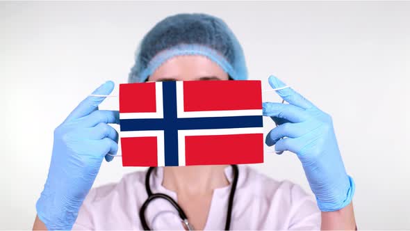 Close-up. Doctor in Glasses, Blue Medical Cap, Gloves Holds in Hands Medical Mask with Norway Flag