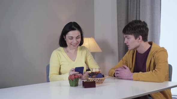 Young Brunette Caucasian Woman Sitting at the Table and Using Phone As Teenage Boy Coming Up 