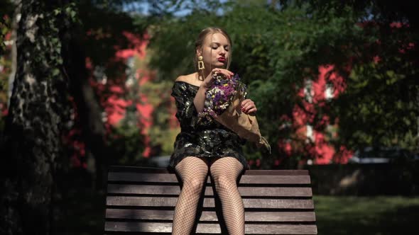 Front View Drunken Laughing Young Woman Sitting on Bench in Sunlight Tearing Flowers Petals