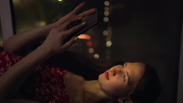 girl holds phone and looks at it at window against background of night city.