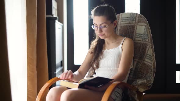 Young girl in glasses reads a book in natural light on a rocking chair