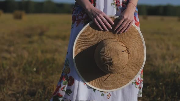 Woman in White Dress Closeup Holds Hat in Her Hands on Background of Field