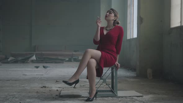 Portrait Elegant Woman in Red Elegant Dress Sitting on the Chair in the Abandoned Building Smoking