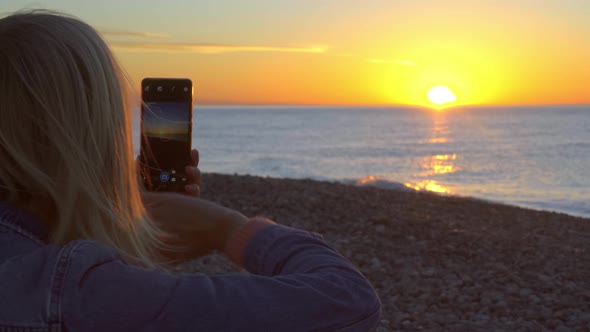 Women's hands hold a smartphone and take pictures of the sunset on the sea.