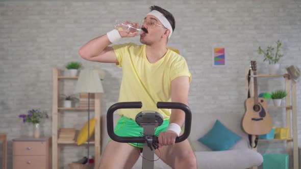 Funny Sportsman From the 80's with a Mustache Is Engaged on a Exercise Bike at Home and Drinks Water