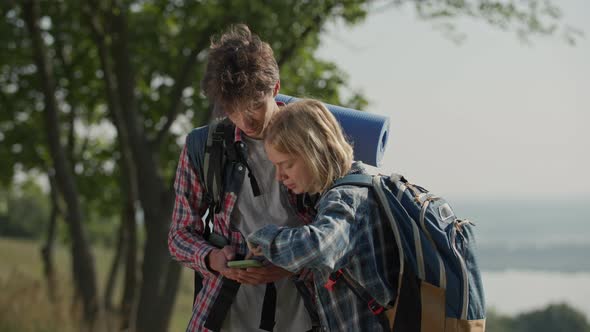 Young Adult Tourist Couple with Travel Backpacks Looking at Mobile Phone for Trail or Route to Hike