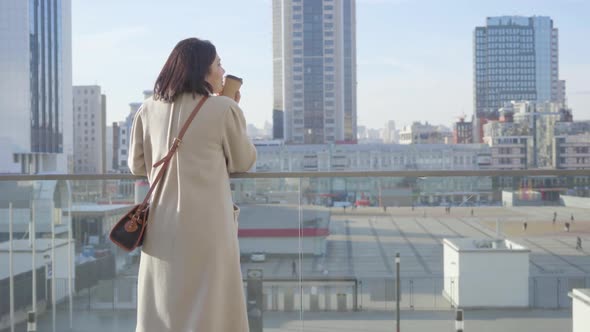 Back View of Young Caucasian Businesswoman Standing with Coffee Cup and Looking at City Street